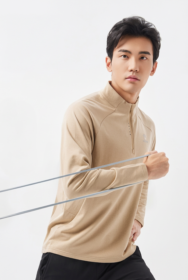 A man wearing a beige long-sleeve athletic quarter-zip pullover and black pants, holding a silver resistance band against a plain white studio background.
